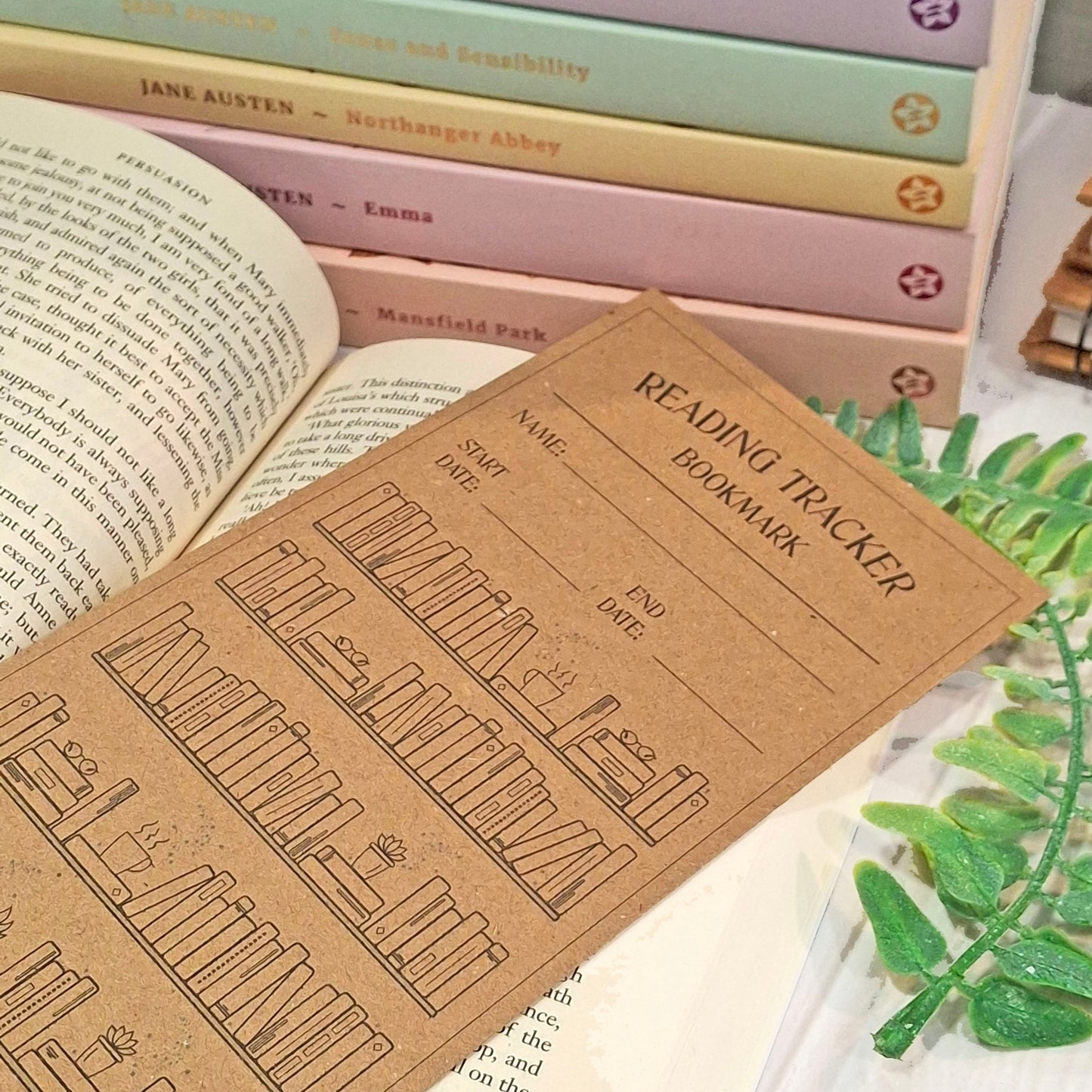 A brown kraft card reading tracker bookmark with black printed bookshelf illustrations and text, laid over open books with a stack of pastel-coloured books in the background and a green leafy sprig on the right.