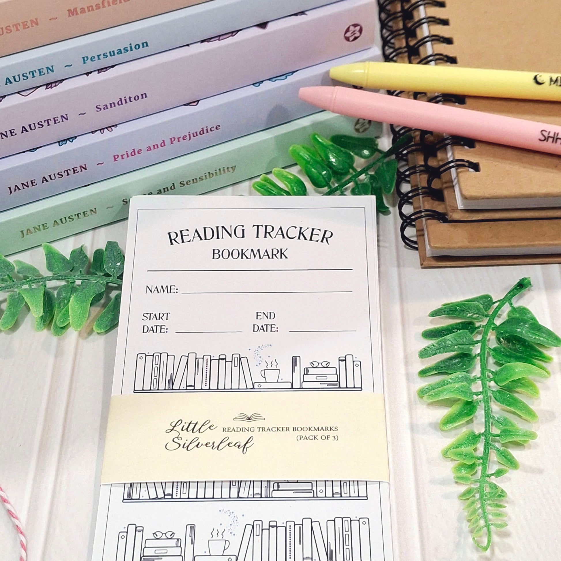 A set of reading tracker bookmarks by Little Silverleaf arranged on a white surface with books, pens, and decorative greenery. The bookmarks are white with black line art of bookshelves and a cream band at the bottom.