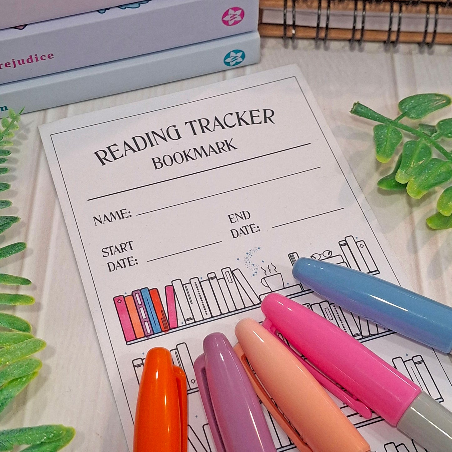 A partly coloured in reading tracker bookmark sheet with book illustrations, styled on a light wood surface with colourful highlighters and small green plant leaves.