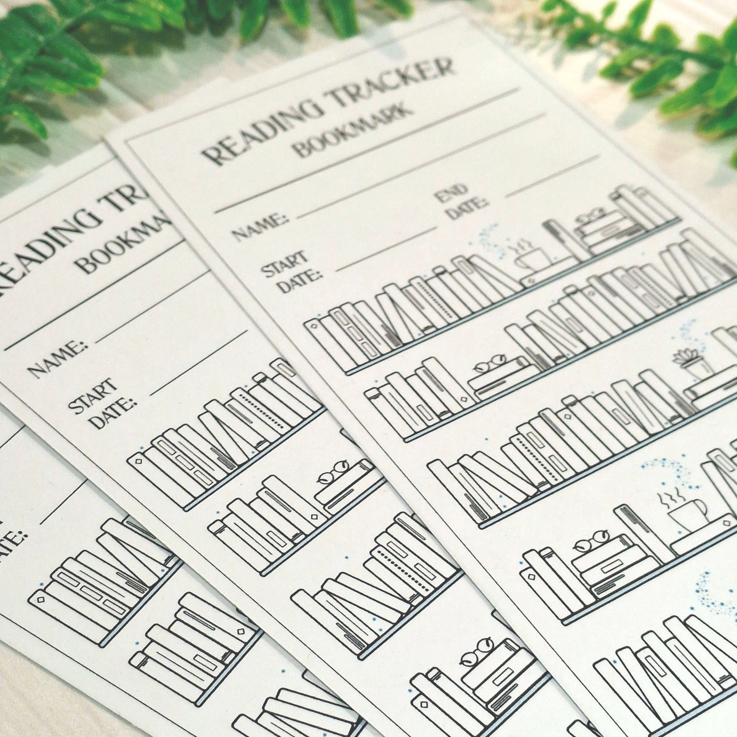 A close-up of several printed reading tracker bookmarks fanned out on a light surface with blurred green leaves in the background. The bookmarks are white with black text and line drawings of book spines and shelves.