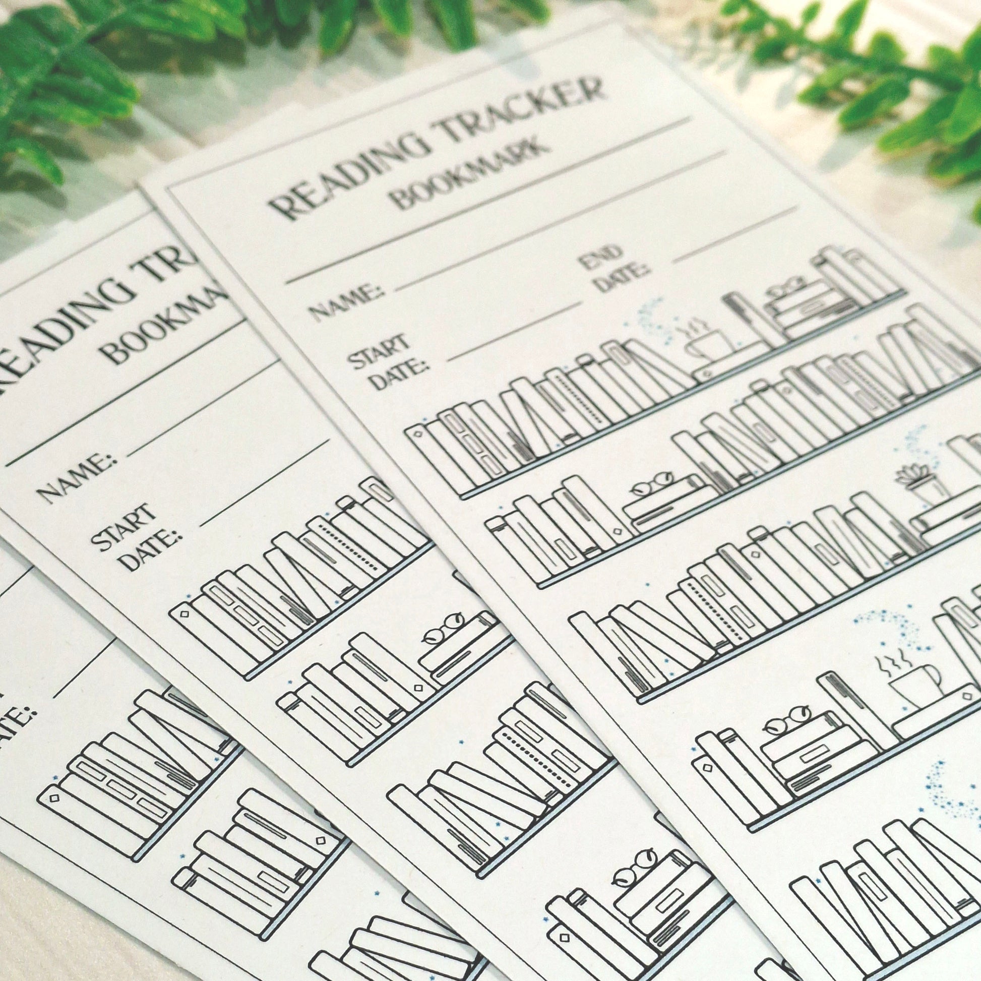 A close-up of several printed reading tracker bookmarks fanned out on a light surface with blurred green leaves in the background. The bookmarks are white with black text and line drawings of book spines and shelves.