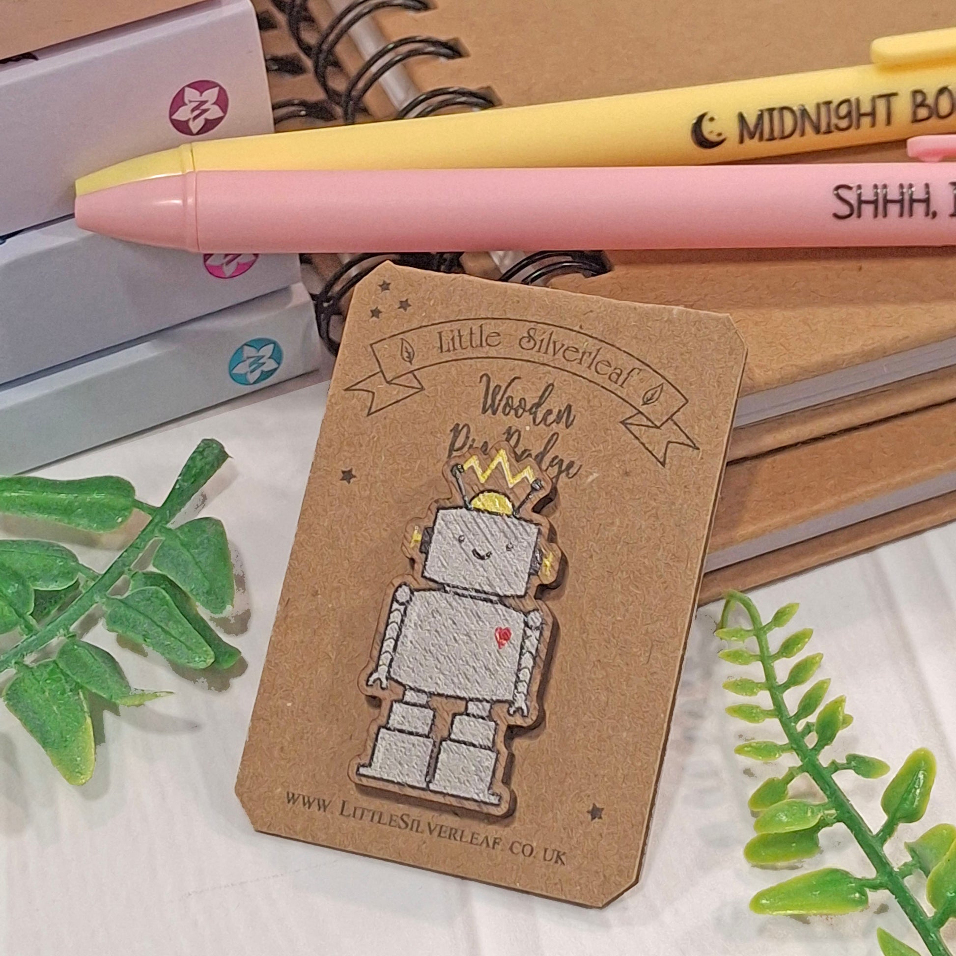 A small robot-shaped pin badge on a brown kraft backing card, styled with pink and yellow pens, notebooks, and green leaves on a light surface.