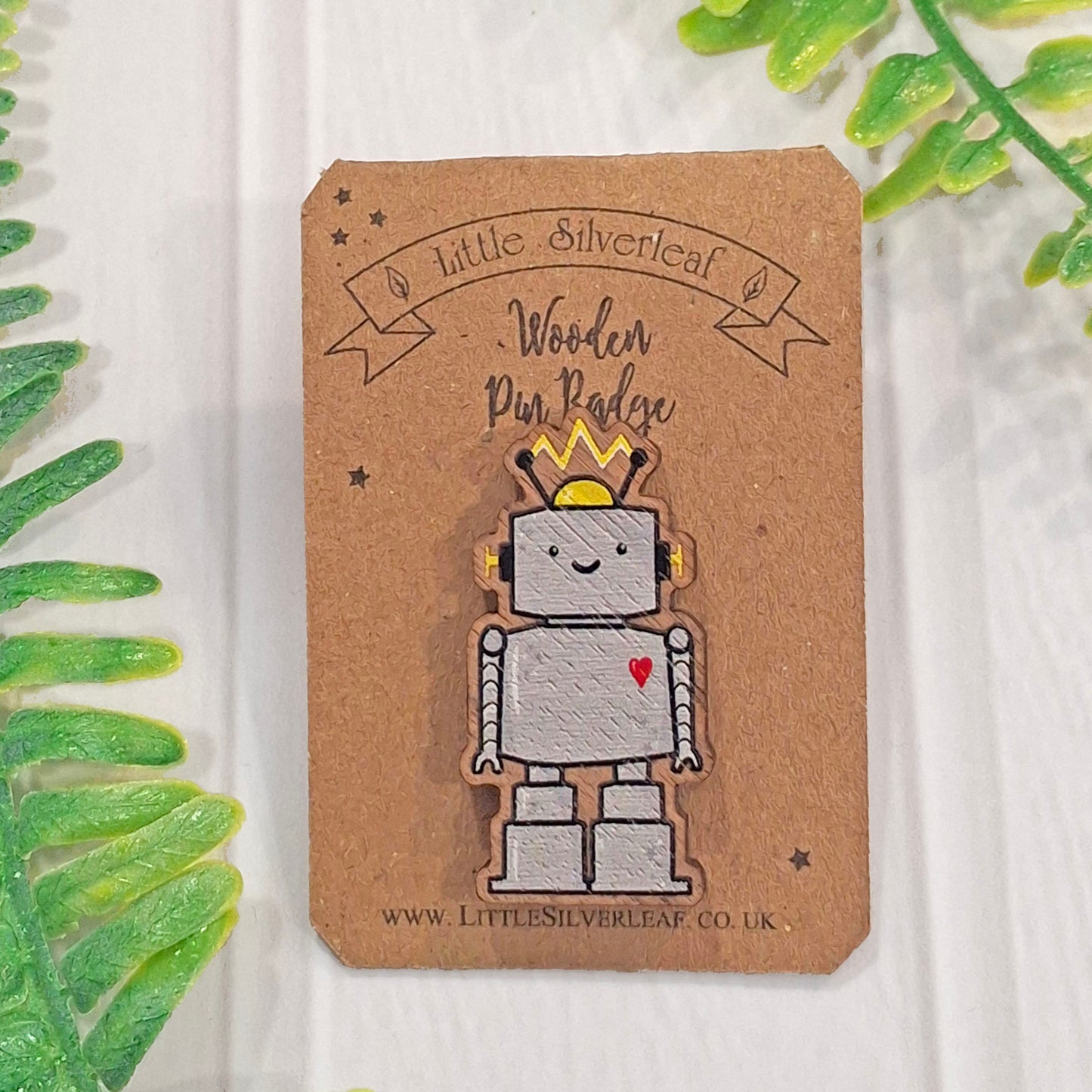 A wooden pin badge shaped like a smiling silver robot with a red heart, mounted on a brown kraft card labeled 'Little Silverleaf Wooden Pin Badge'.