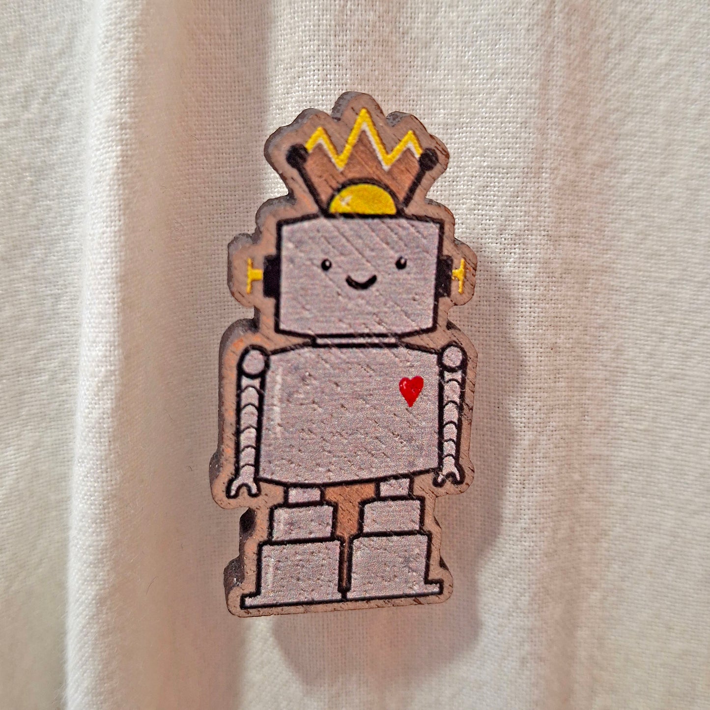 A wooden robot pin badge with a smiling face and red heart, shown against a light beige fabric tote bag background.