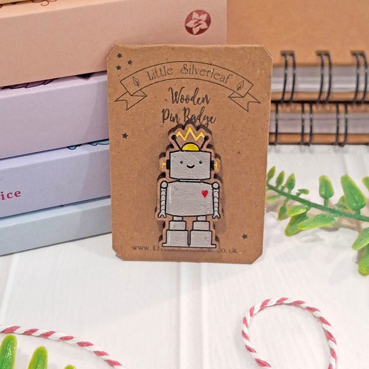 A robot pin badge on a brown kraft backing card, photographed on a white surface with notebooks, greenery, and bakers twine in the background.