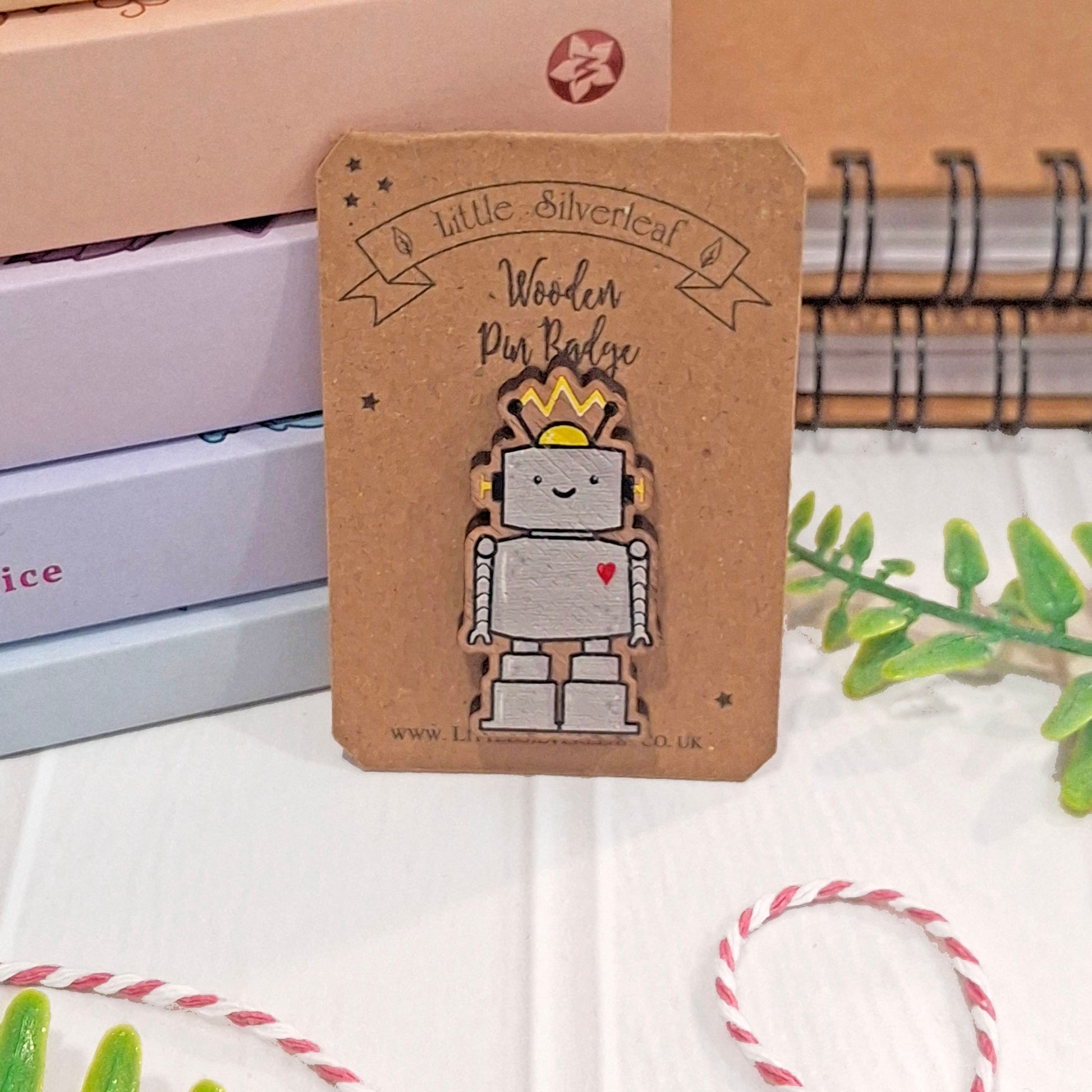 A robot pin badge on a brown kraft backing card, photographed on a white surface with notebooks, greenery, and bakers twine in the background.