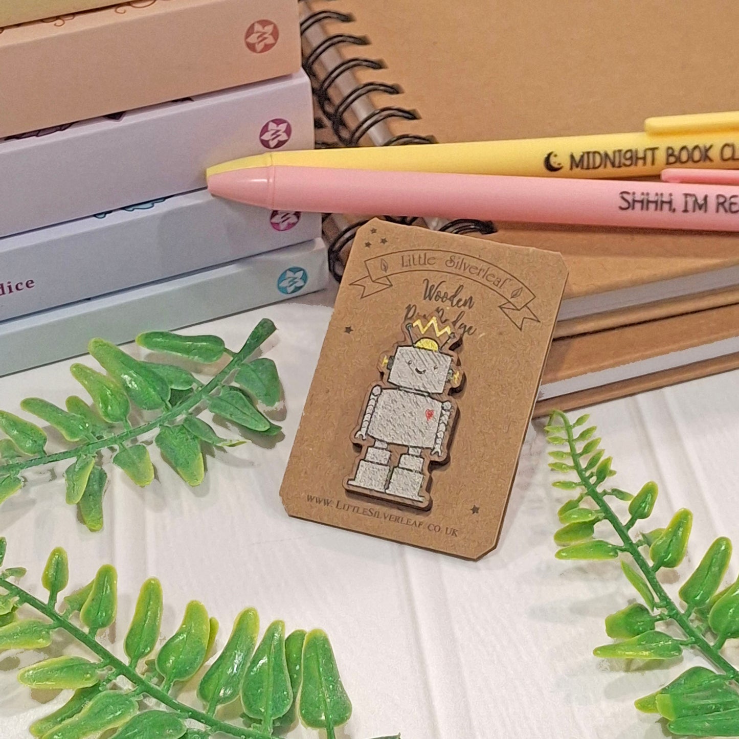 A small robot pin on a kraft display card, styled on a white surface with books, a yellow pen, and green leaves around it.