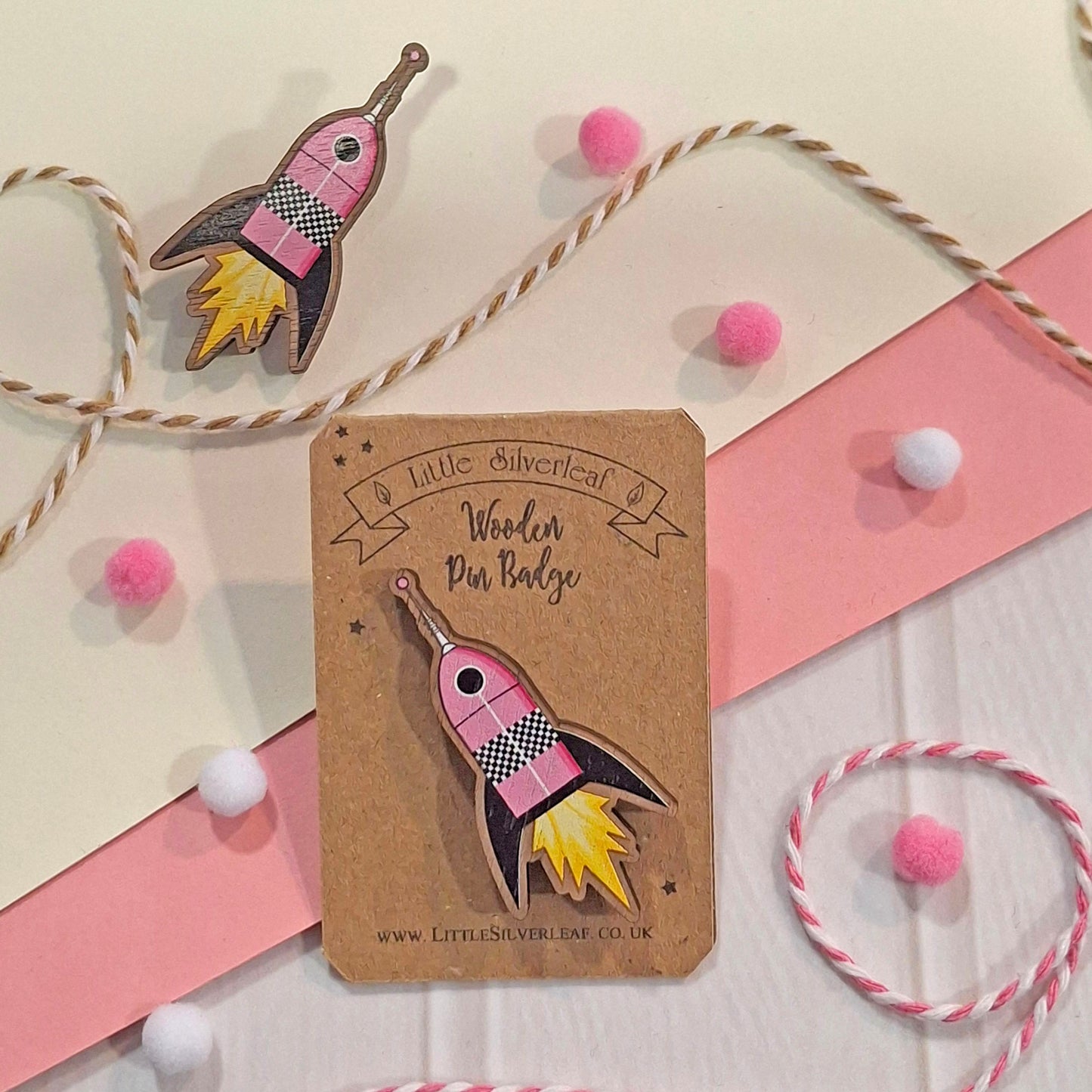 A pink and yellow rocket-shaped wood pin displayed on a kraft card labelled 'Little Silverleaf Wooden Pin Badge,' with additional matching decorative props including pom-poms, twine, and a second rocket pin nearby.