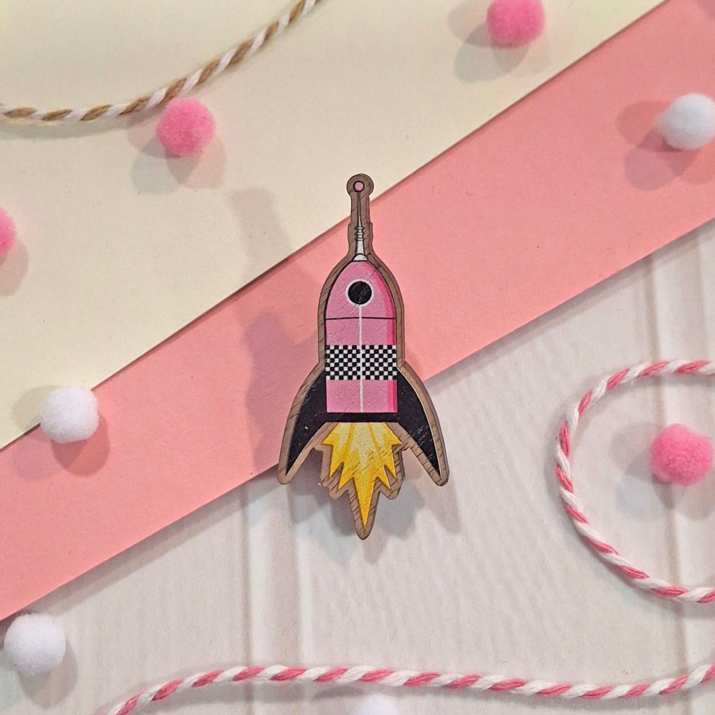 A rocket-shaped wooden pin with pink, black, and yellow details, displayed on a styled flat-lay background of pink and cream paper with decorative pom-poms and string.