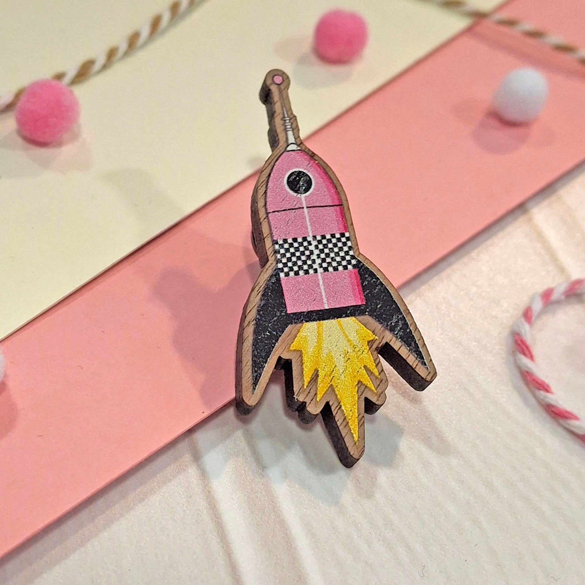 A small wooden rocket-shaped pin badge with pink, black, yellow, and white details, photographed on a pastel card with pink and cream sections and small pom-pom decorations.