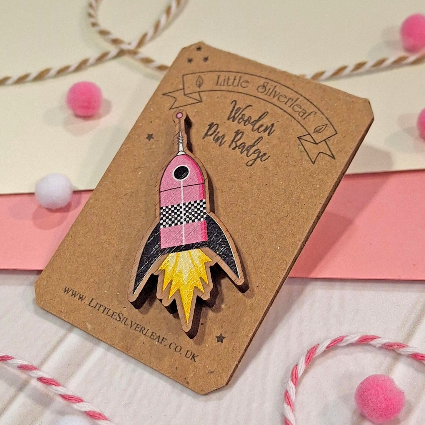 A wooden pin badge shaped like a pink and black rocket ship with yellow flames, mounted on a brown kraft card labelled 'Little Silverleaf Wooden Pin Badge.' The card is styled on a light surface with pink and white pom-poms and twisted twine.