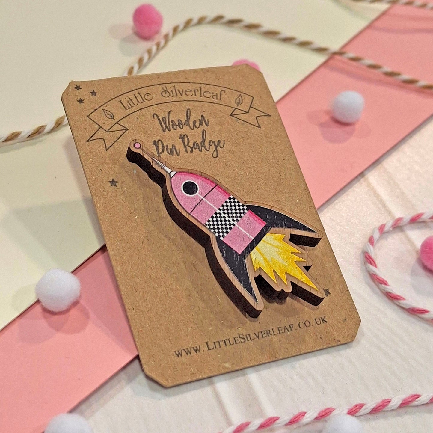 A wooden pin badge shaped like a pink rocket ship with black and yellow flame details, mounted on a brown kraft card from Little Silverleaf. The card is styled on a pink and cream background with decorative pom-poms and twine.