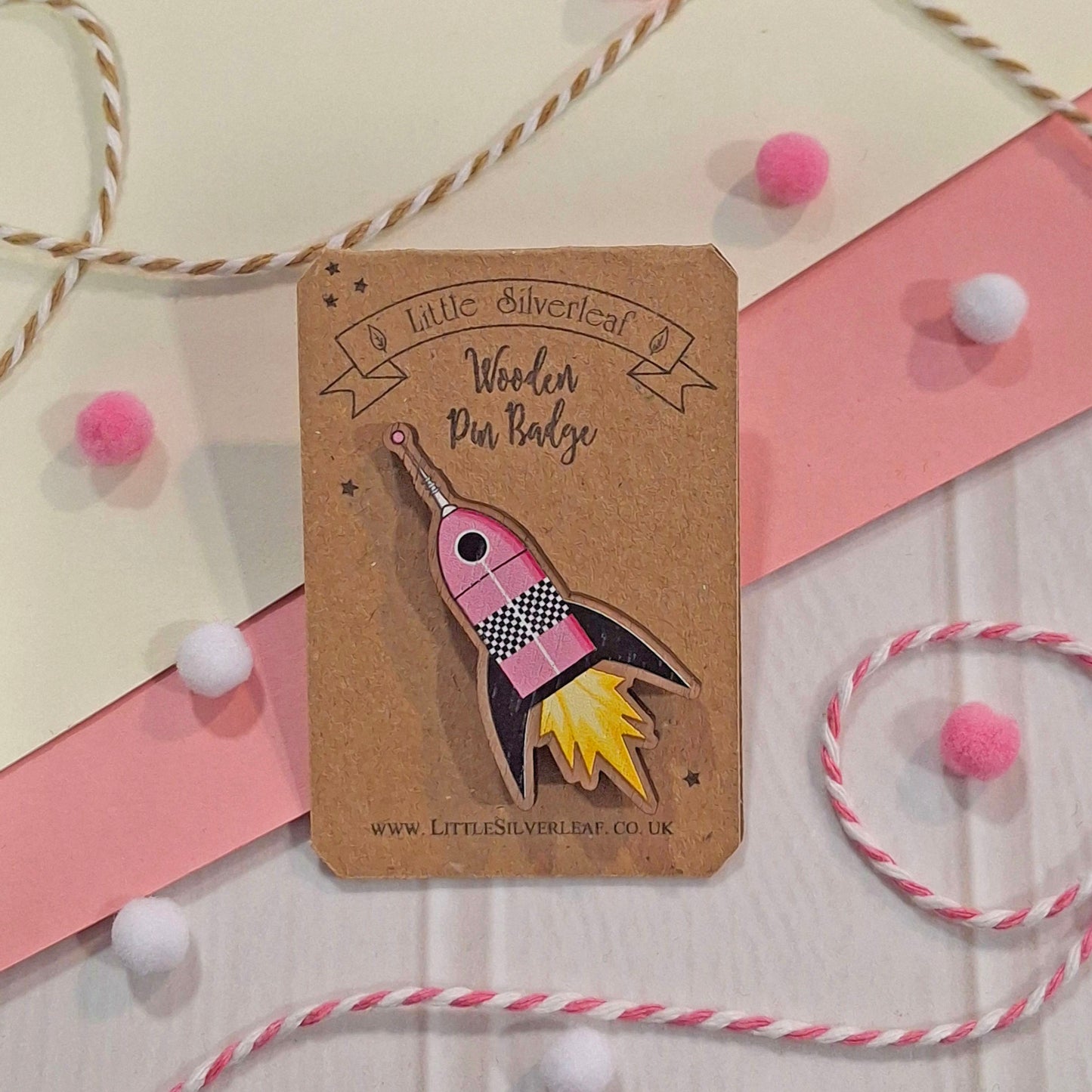 A wooden pin badge shaped like a pink rocket ship with black, white, and yellow details, mounted on a brown kraft card. The card is photographed on a styled flat-lay background with pink and cream paper borders, pink and white pom-poms, and twisted string.