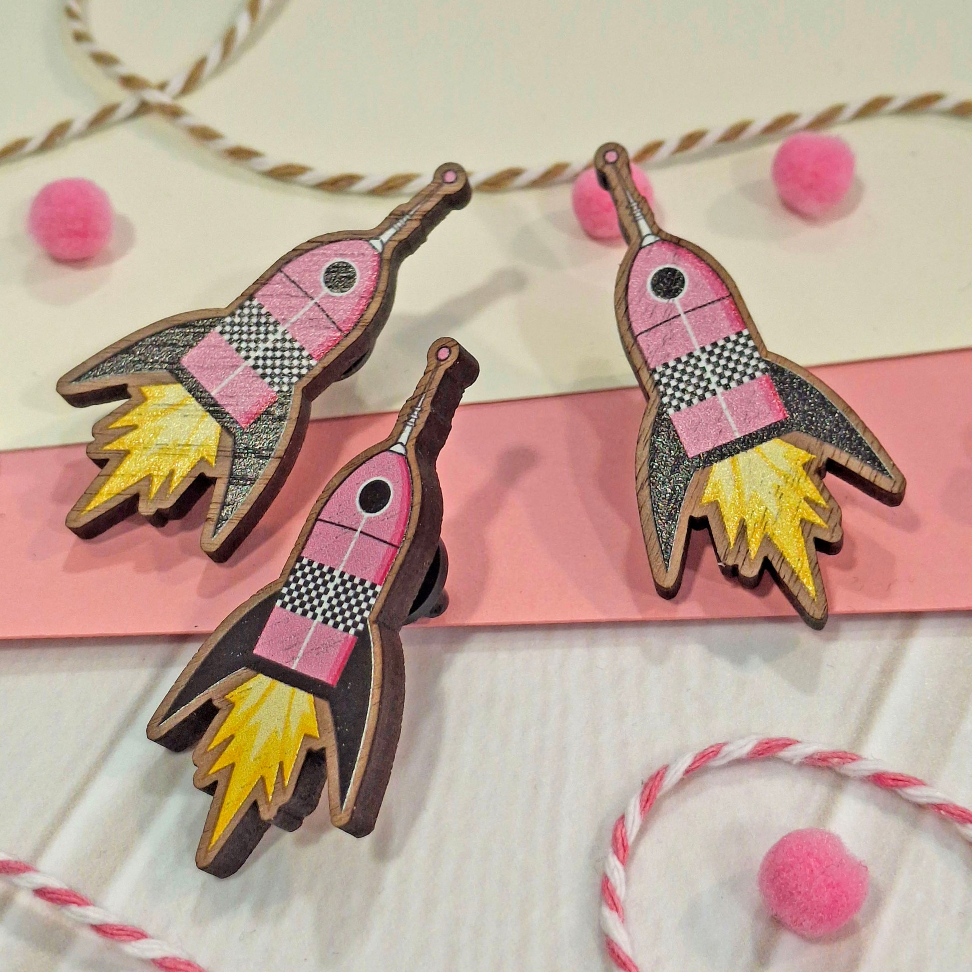 Three hand-painted wooden rocket-shaped pin badges with pink, black, yellow, and white details, displayed on a pink and white striped card with decorative pink pom-poms and twine.