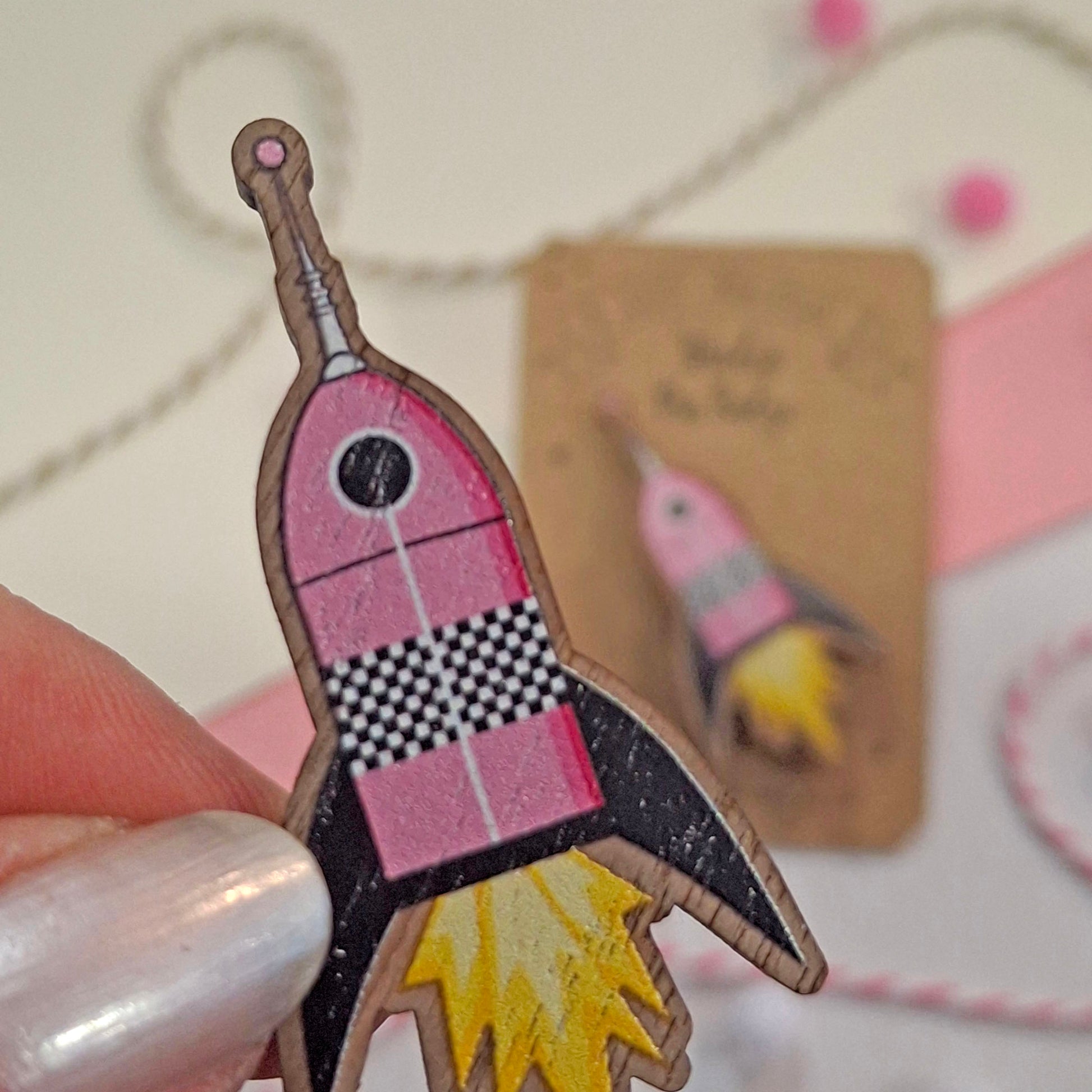 A hand holds a small pink rocket-shaped wooden pin badge with a black-and-white checkered section and yellow flame details. A similar item is blurred in the background on a kraft card, with pastel paper and twine around it.