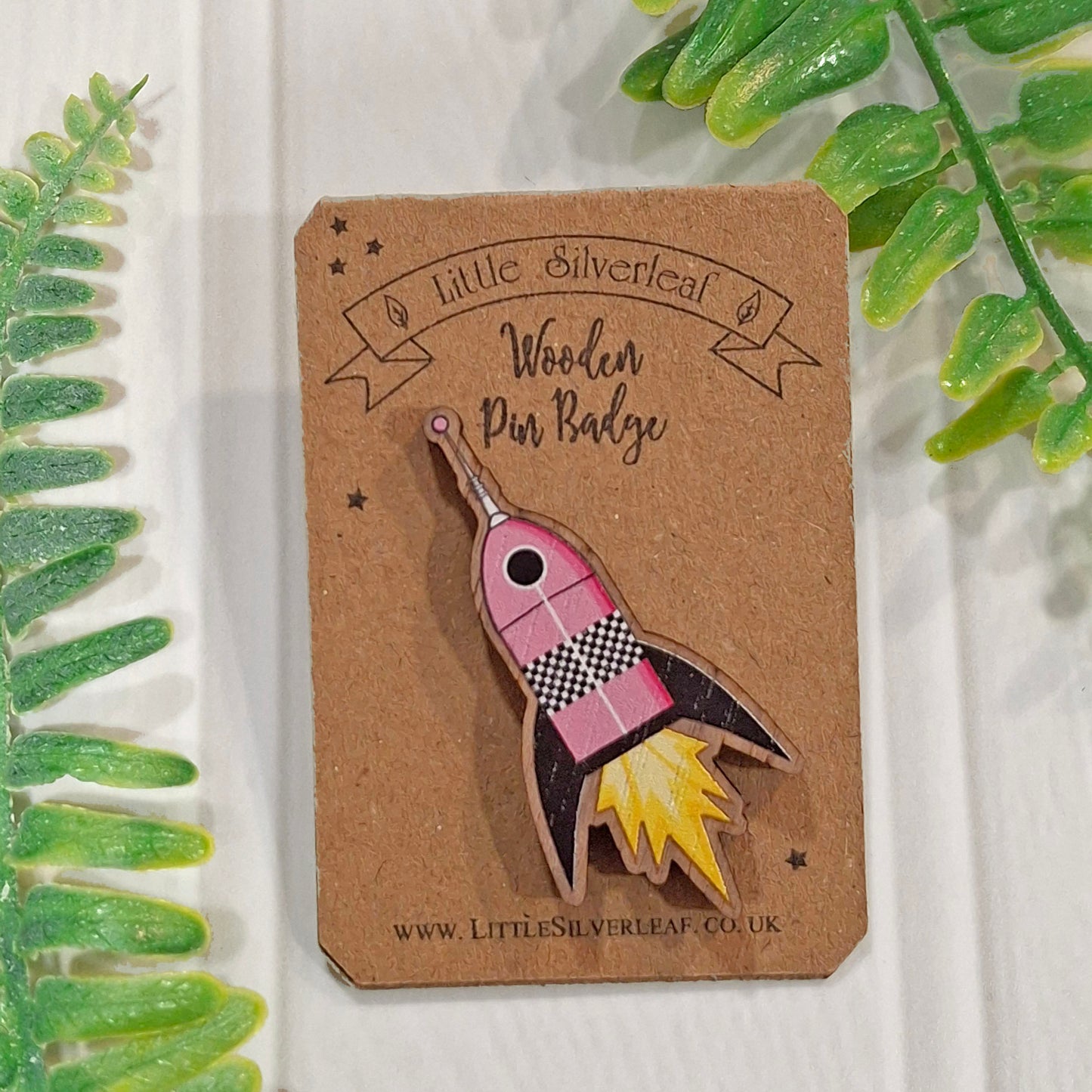 A wooden pin badge shaped like a pink and black rocket ship with yellow flames, mounted on a brown kraft card labeled 'Little Silverleaf Wooden Pin Badge'. The card is photographed on a white textured surface with green leaves framing the sides.