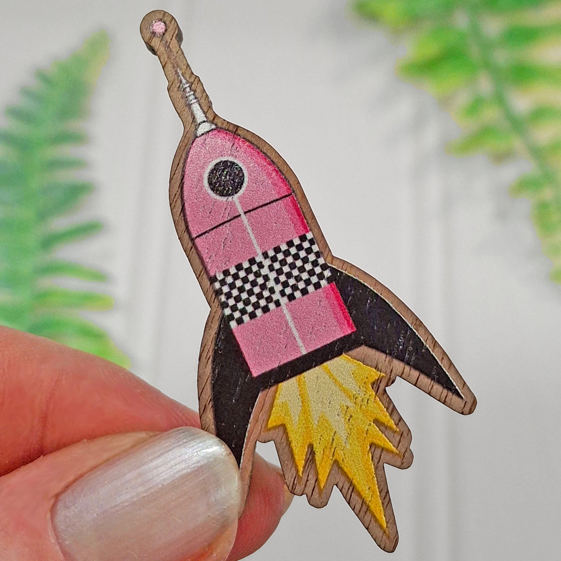 A wooden rocket-shaped pin badge with pink and black sections and a chequered pattern and yellow flame details, being held between fingers, and set against a softly blurred background with green leaves.