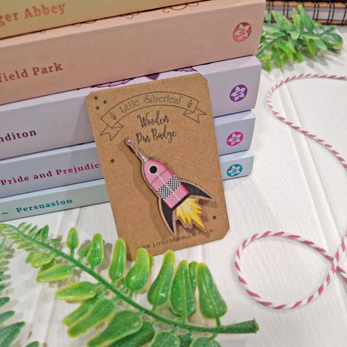 A small pink and black rocket-shaped pin badge on a brown kraft backing card, styled on a white surface with stacked books, green leaves, and pink-and-white twine.