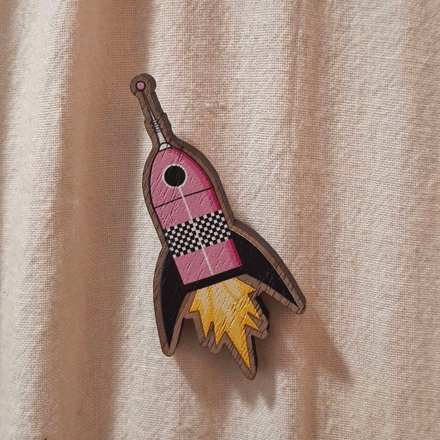 A wooden rocket pin badge on a cream fabric tote bag background.