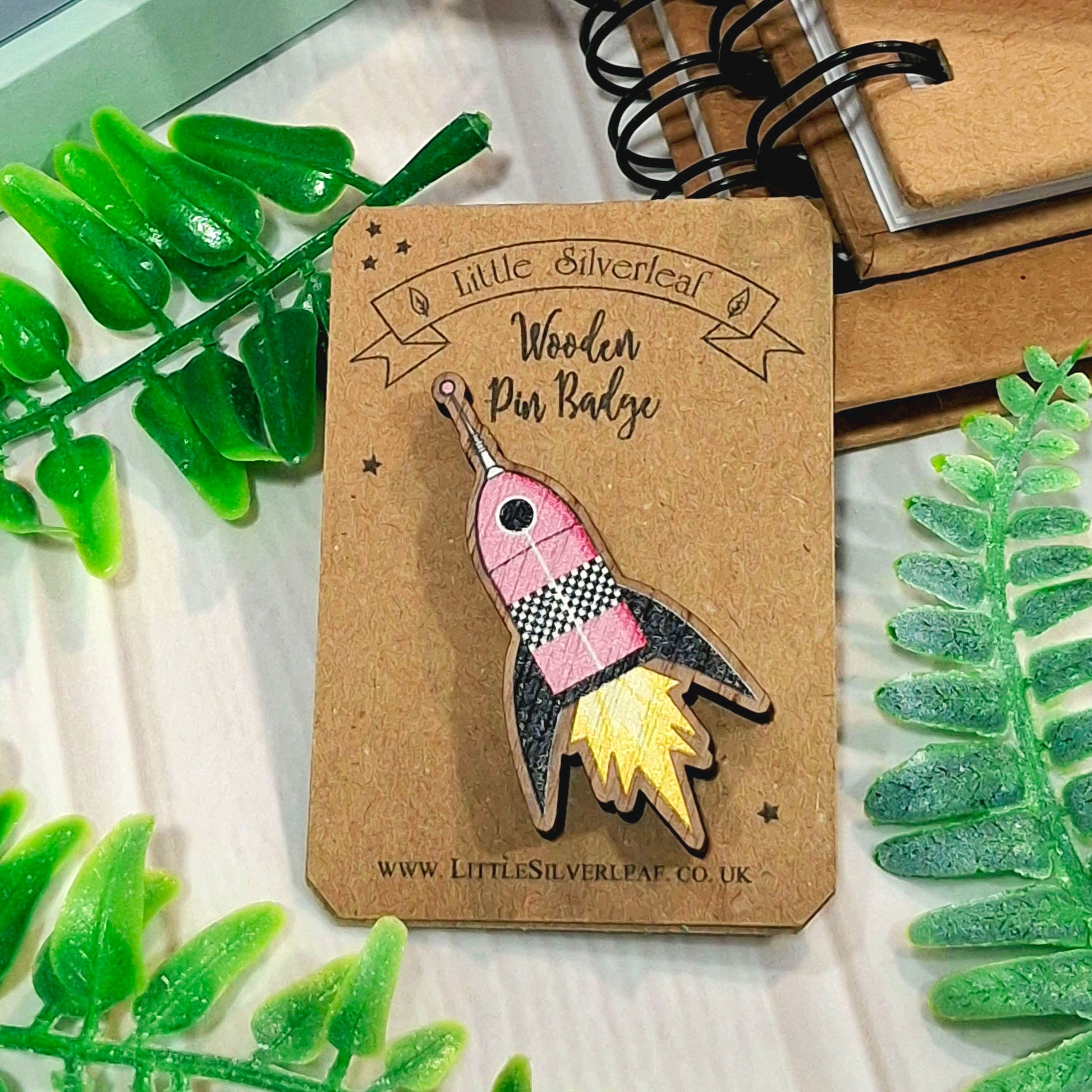 A wooden pin badge shaped like a pink and black space rocket ship with yellow flames, mounted on a brown kraft backing card. The card is styled with green leaves and a spiral notebook on a light surface.