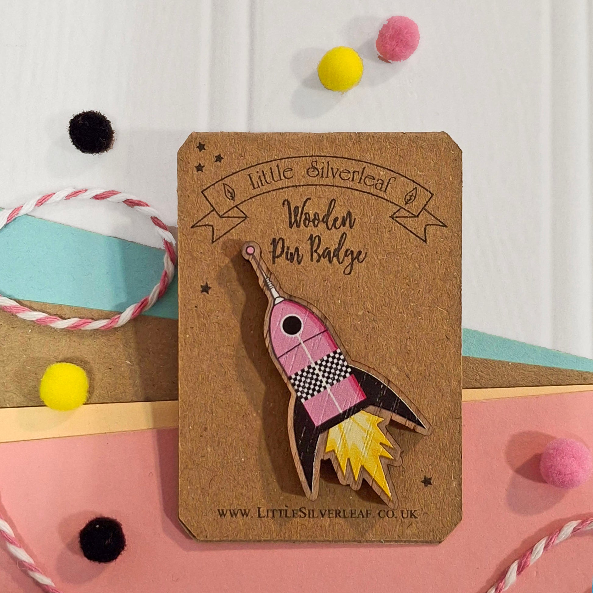 A wooden pin badge shaped like a pink rocket ship with yellow flames, mounted on a brown kraft display card. The card is labelled 'Little Silverleaf Wooden Pin Badge'.