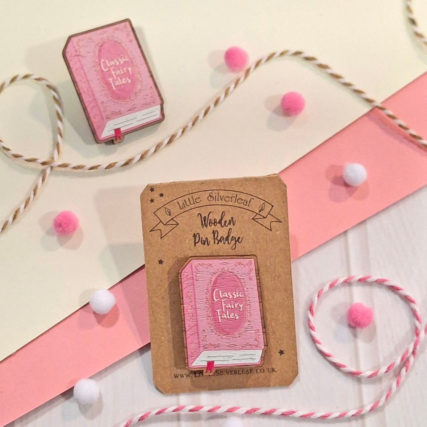 A pink wooden pin shaped like a classic fairy tale book, mounted on a kraft display card. The pin is styled on a light cream background with pink paper, bakers twine, and small pink and white pom-poms.