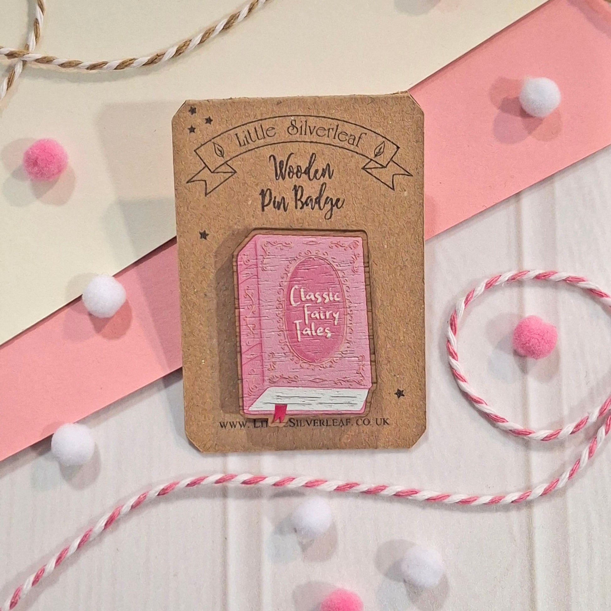 A wooden pin badge featuring a pink book illustration labelled 'Classic Fairy Tales,' mounted on a kraft card. The badge is photographed on a light surface with pink ribbon, white pom-poms, and pink-and-white string as styled props.