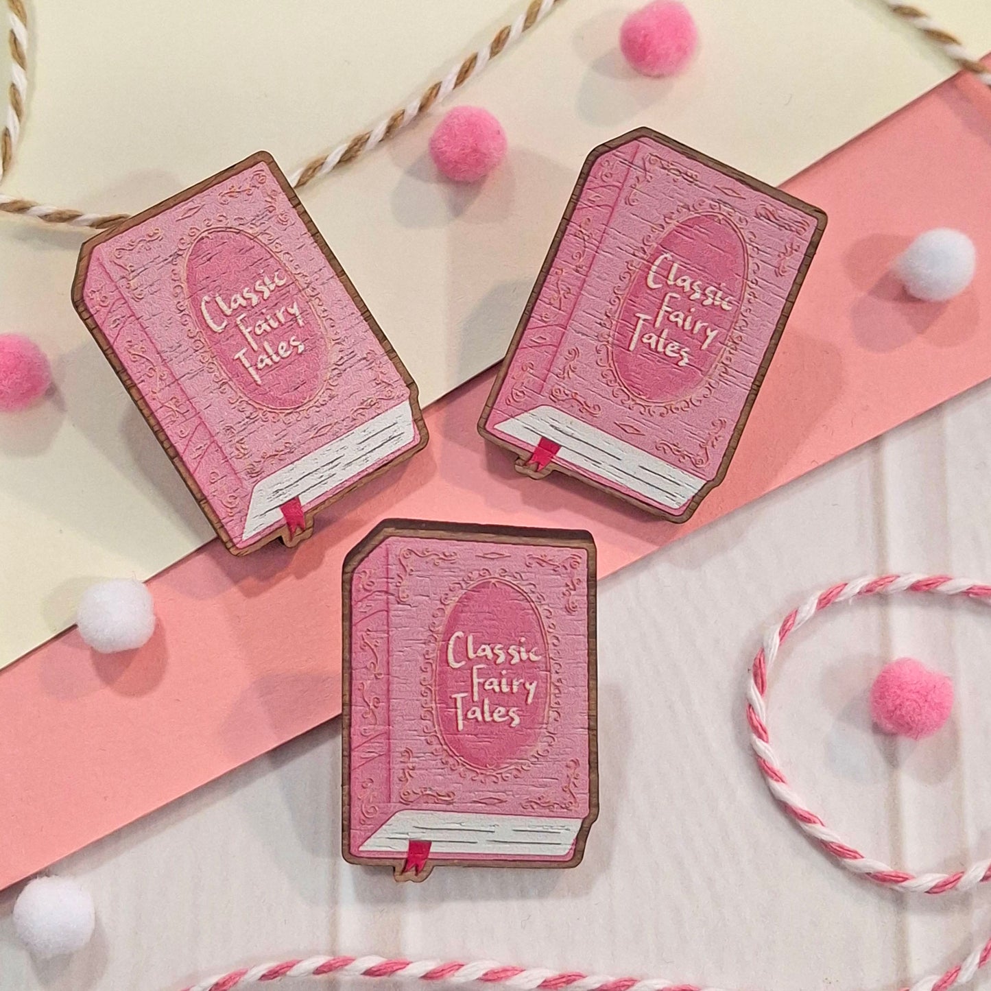 Three pink book-shaped pins with 'Classic Fairy Tales' on the cover, arranged on a pink and cream flat-lay background with decorative pom-poms and ribbon.