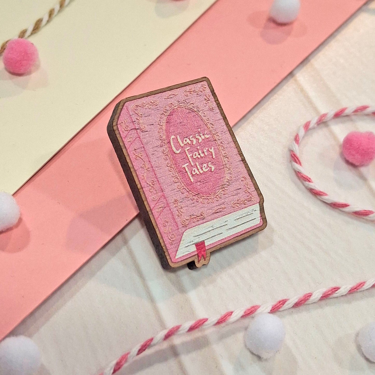 A book-shaped wooden pin badge that reads 'Classic Fairy Tales' in pink, styled on a white surface with pink paper, pom-poms, and twisted string.