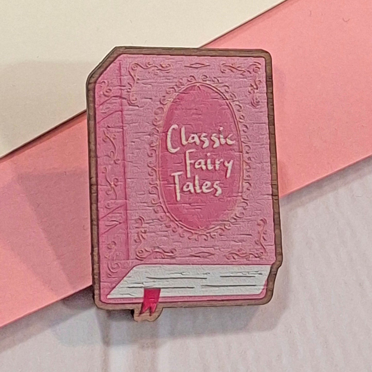 A pink wooden pin shaped like a vintage book titled 'Classic Fairy Tales', displayed on a pink and white background.