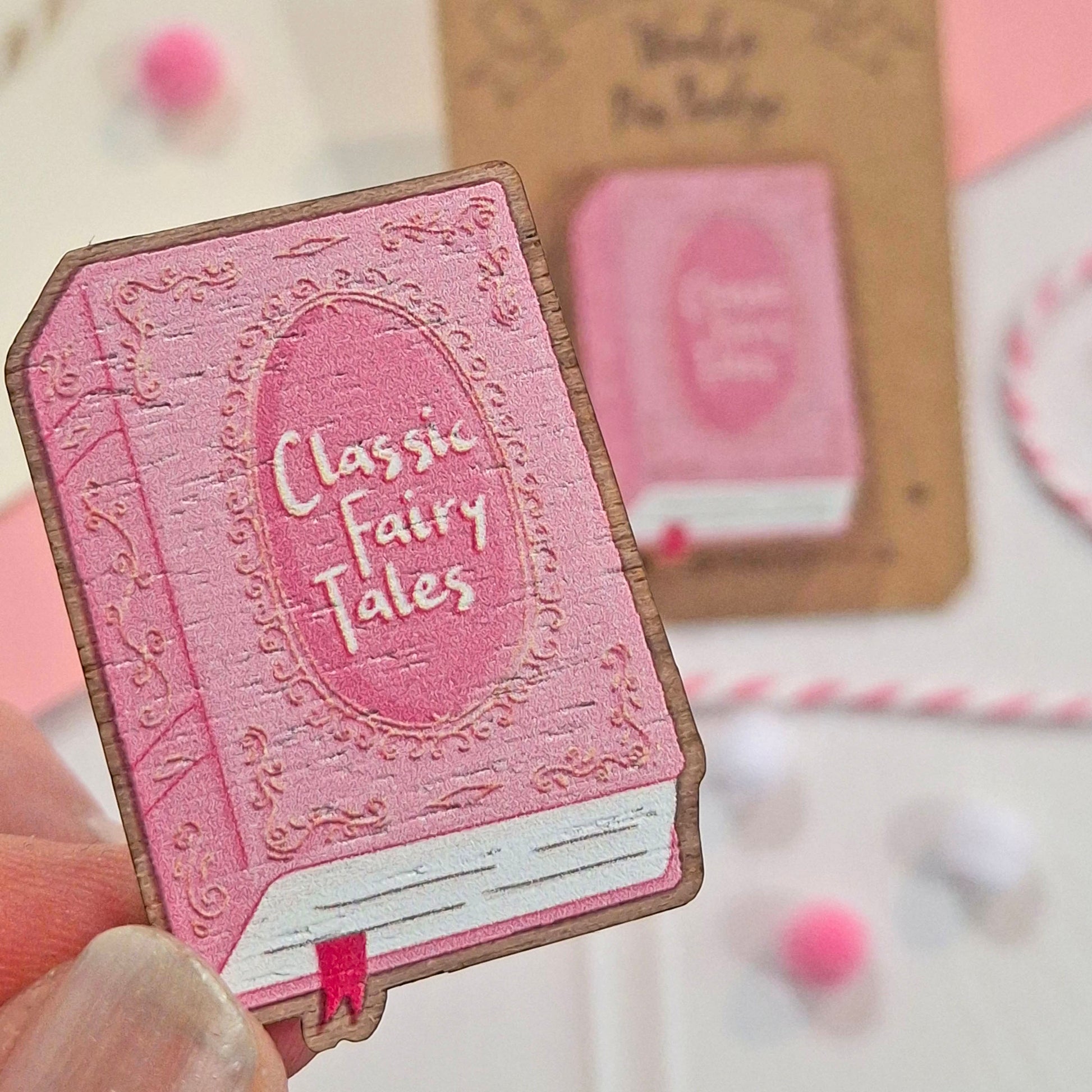 A hand holds a small pink book-shaped pin badge that reads 'Classic Fairy Tales' in front of a similar blurred item on a card. The scene is styled with pastel colours and decorative paper elements.