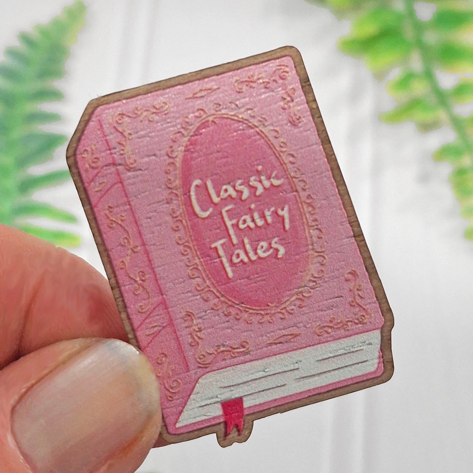 A hand holding a pink book-shaped pin badge with decorative details and the words 'Classic Fairy Tales' on the front, against a softly blurred white background with green leaves.