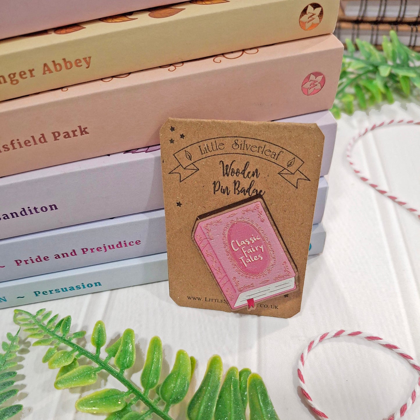 A wooden pin badge shaped like a pink classic fairy tale book, mounted on a brown kraft card. The card is styled on top of a stack of books, with decorative green leaves and pink-and-white string twine on a white wooden surface.