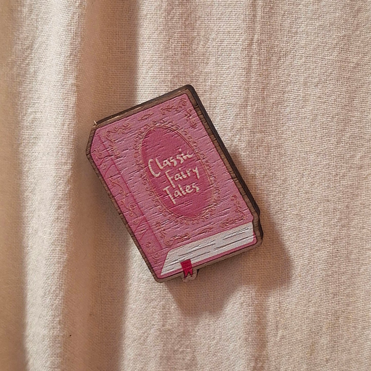 A pink book-shaped badge with 'Classic Fairy Tales' on the cover, resting on a textured beige fabric tote bag background.