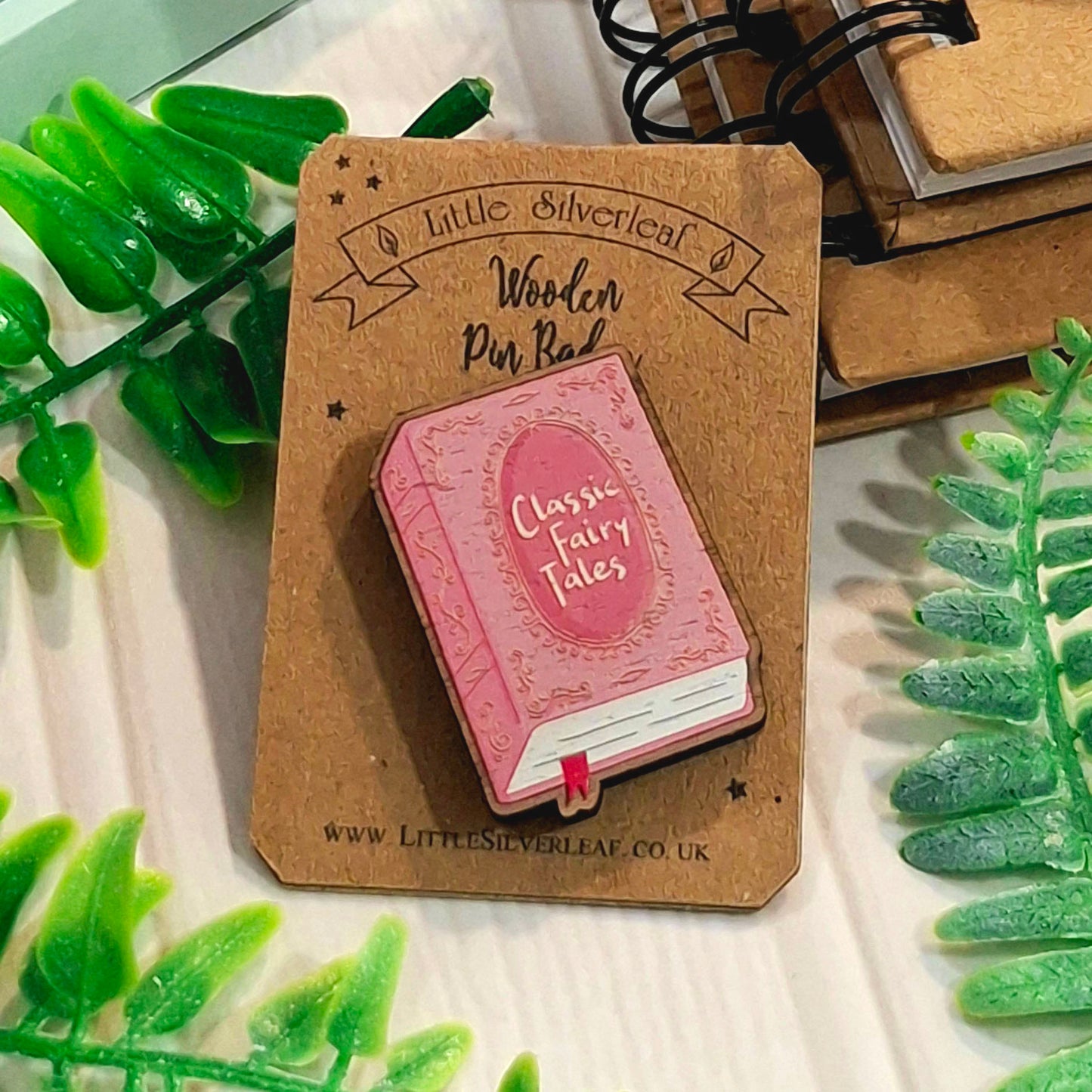 A pink wooden pin shaped like a classic fairy tale book with the words 'Classic Fairy Tales' on the cover, mounted on a brown cardboard display card. The card is labeled 'Little Silverleaf Wooden Pin Badge' and photographed on a light surface with decorative green leaves around it.
