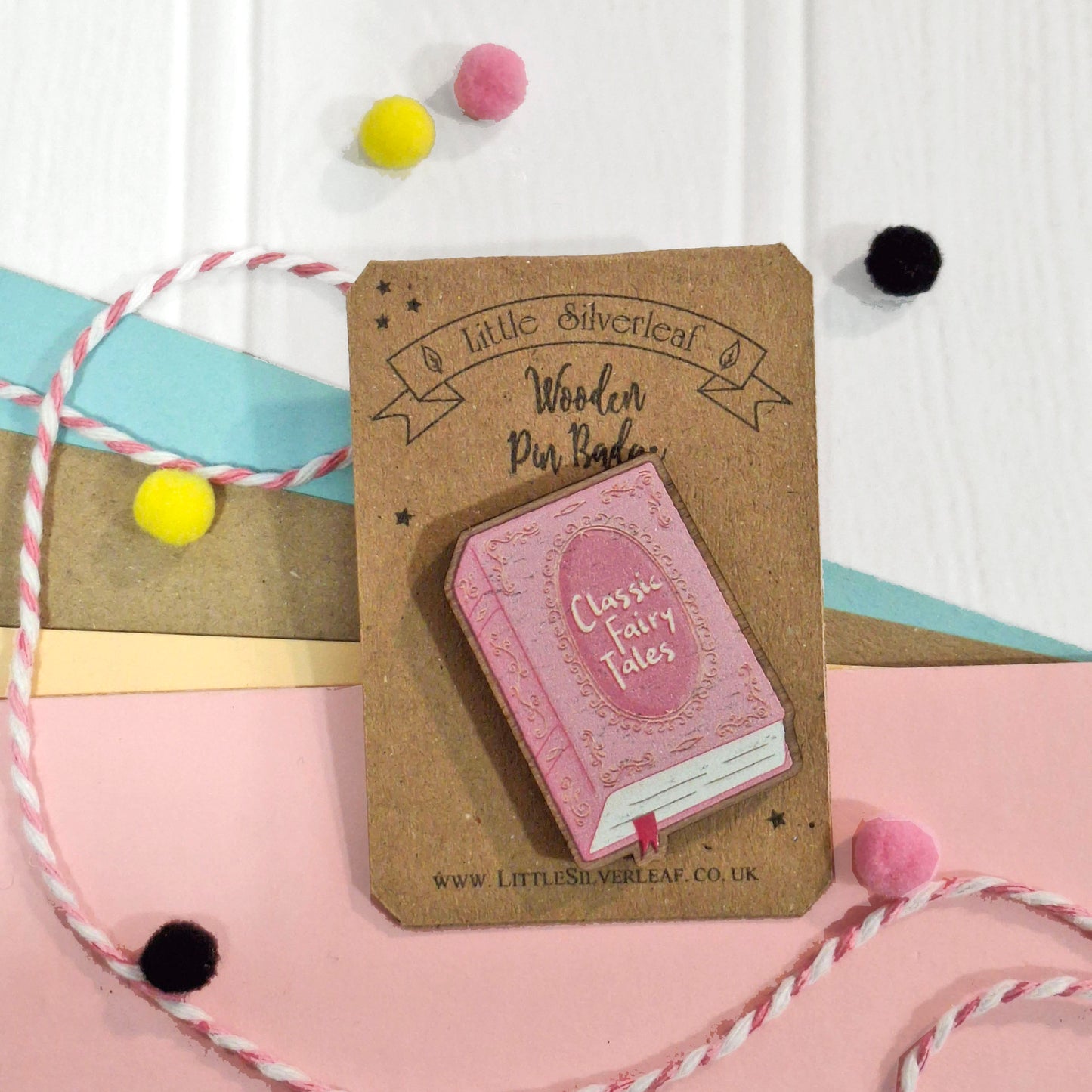 A pink wooden book pin badge on a brown cardboard backing card, styled with pastel paper, bakers twine, and colourful pom-poms.