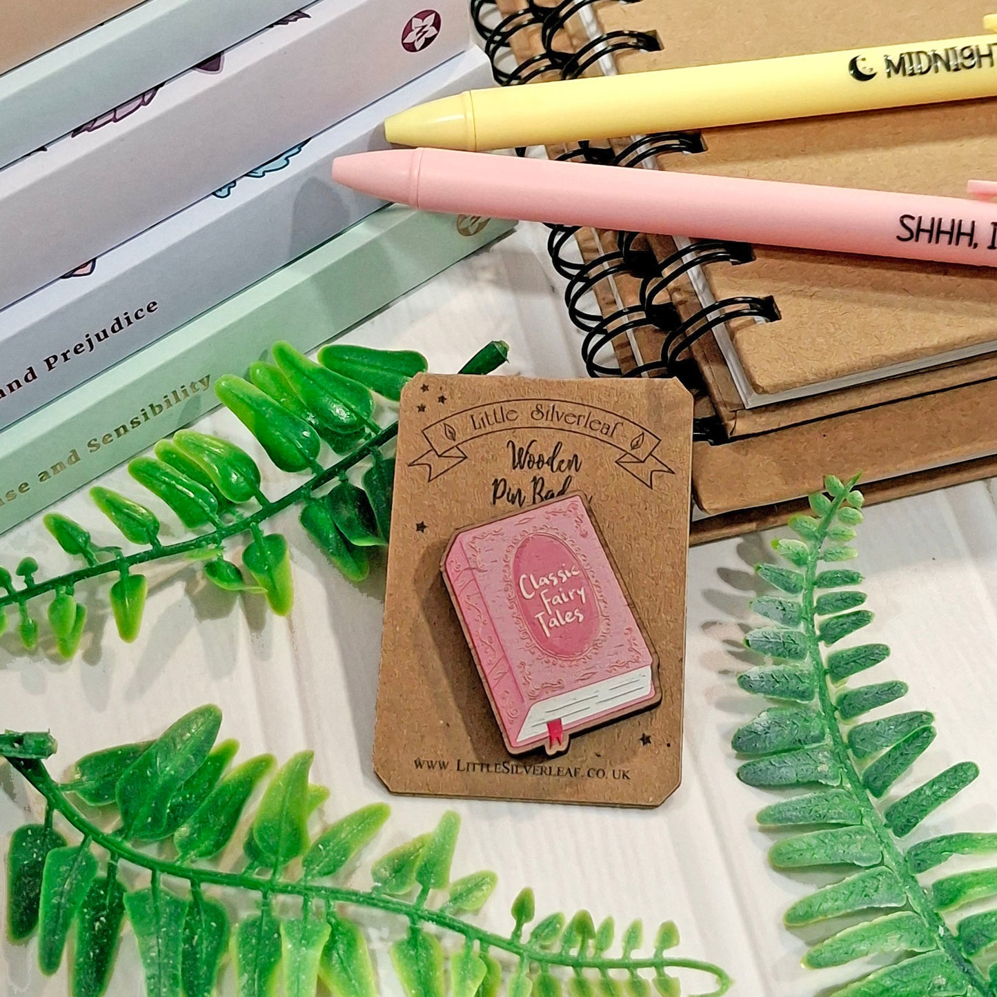 A small pink illustrated book-shaped pin badge displayed on a kraft card, surrounded by notebooks, pens, and green leaf props on a white surface. 